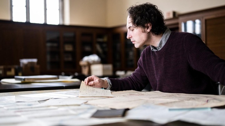 Student sits at a table reading archival documents with boxes and bookshelves in the background